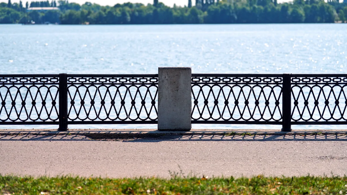 A steel border fence runs beside a wide river under a pale morning sky.