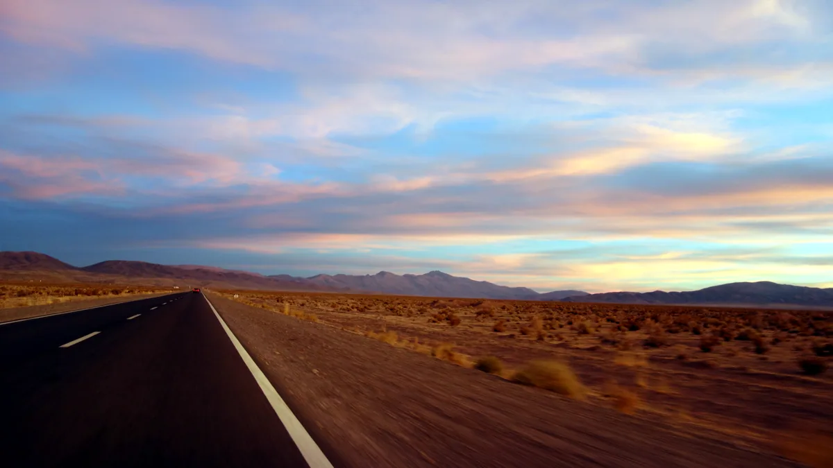 A straight highway crosses open desert as the sun sinks into the haze.