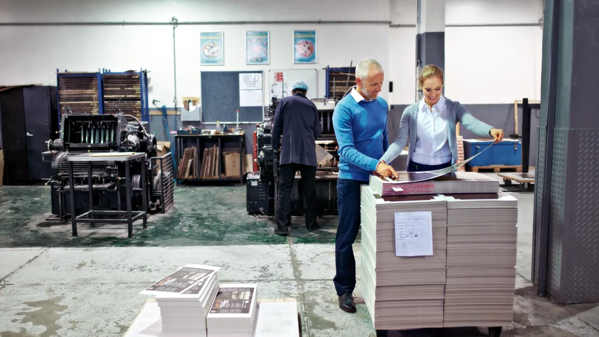 A printing press feeds sheets through a dim newsroom lit by lamp light.